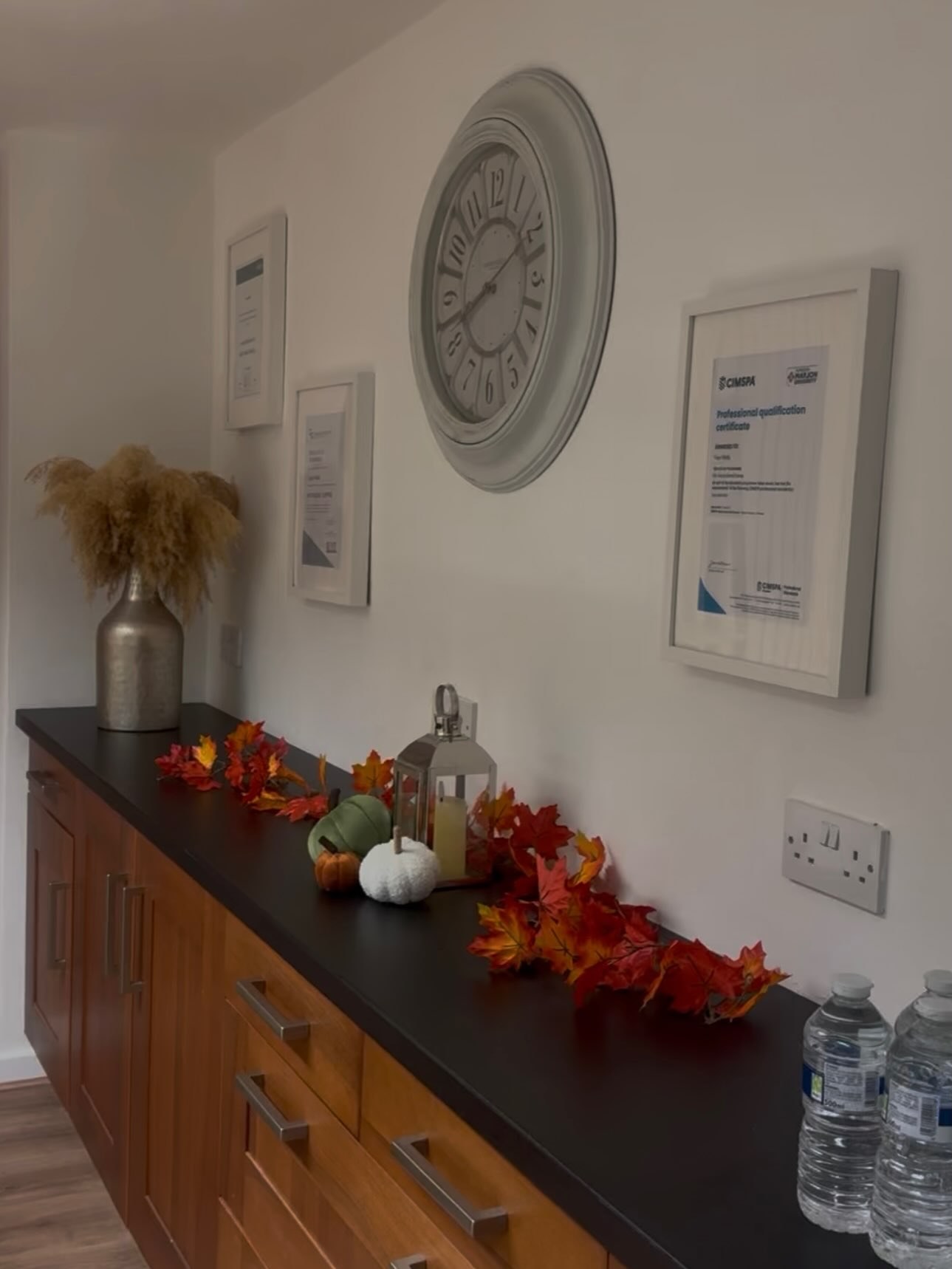 A sideboard with autumn leaves, framed certificates and a large clock at Top Barn